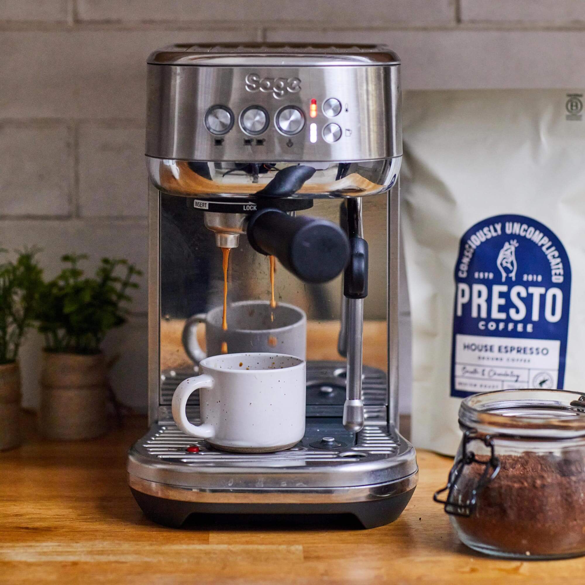 Sage espresso machine brewing coffee into white mug, Presto coffee bag and jar of ground coffee on wooden countertop