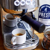 Espresso machine brewing coffee with a freshly poured espresso shot in a white ceramic cup, Prestige coffee bag in the background, ground coffee jar on wooden table.