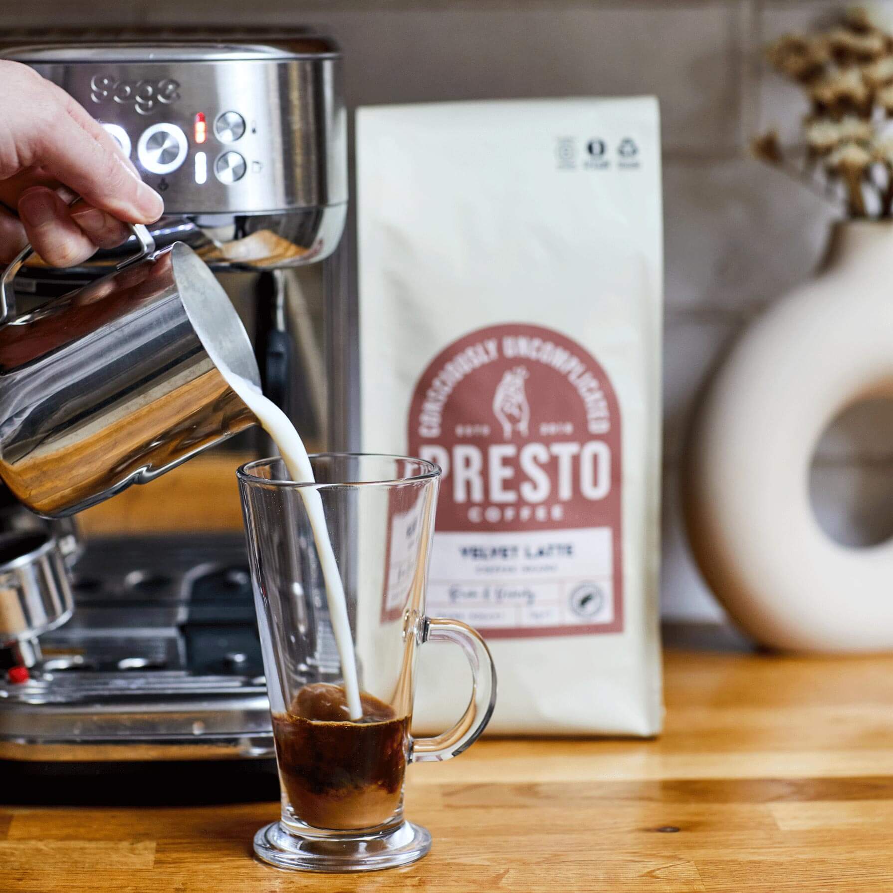 Making a latte with an espresso machine, pouring steamed milk into a glass with espresso, Presto coffee bag on wooden countertop.