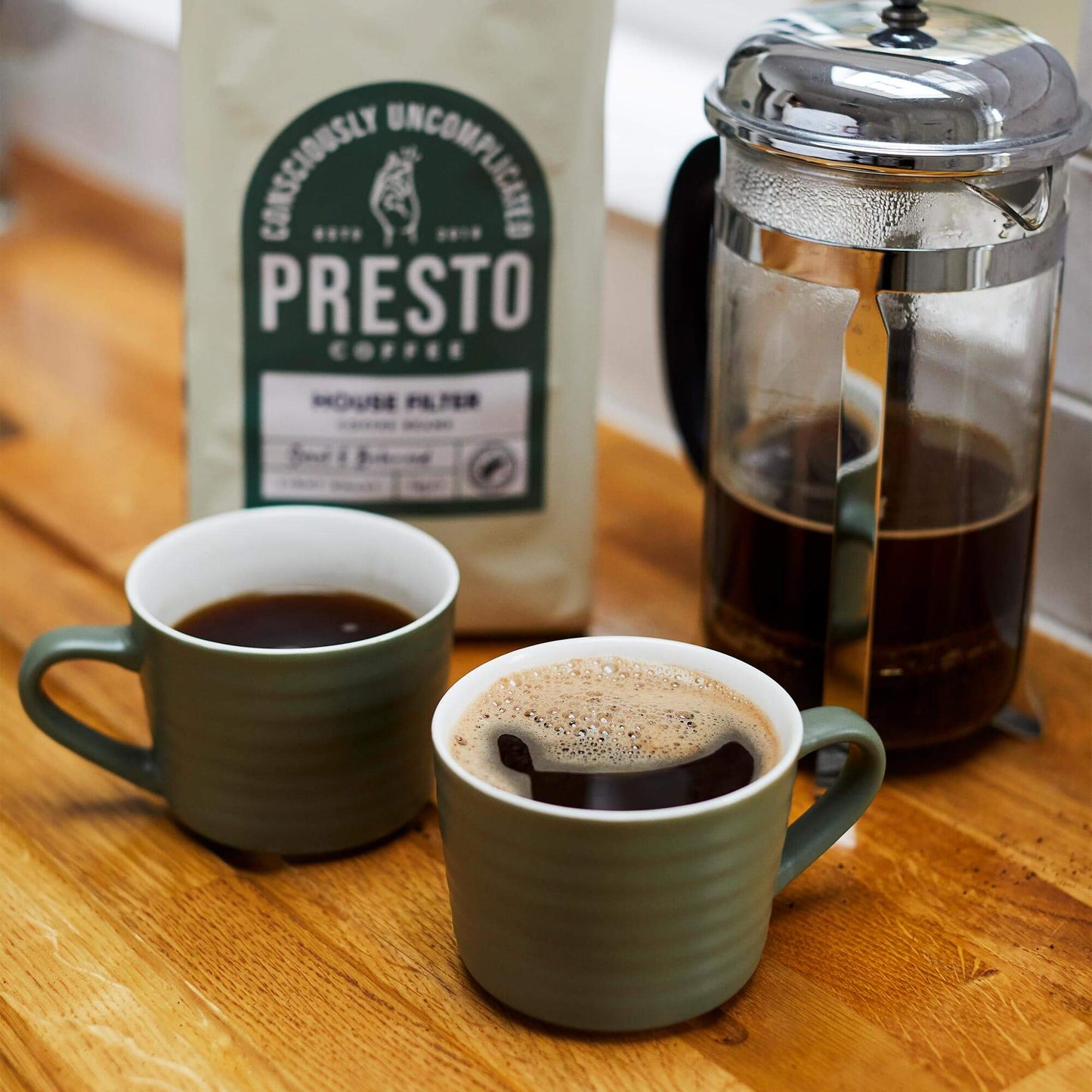 Two green ceramic mugs of freshly brewed coffee next to a French press and a bag of Presto coffee on a wooden kitchen counter.