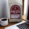 Presto Coffee Intense Espresso beans bag next to a white cup of coffee and a laptop on a wooden table.