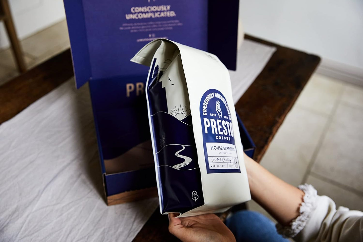 Person holding Presto Coffee bag with blue packaging design, featuring House Espresso medium roast, inside a branded box on a wooden table.