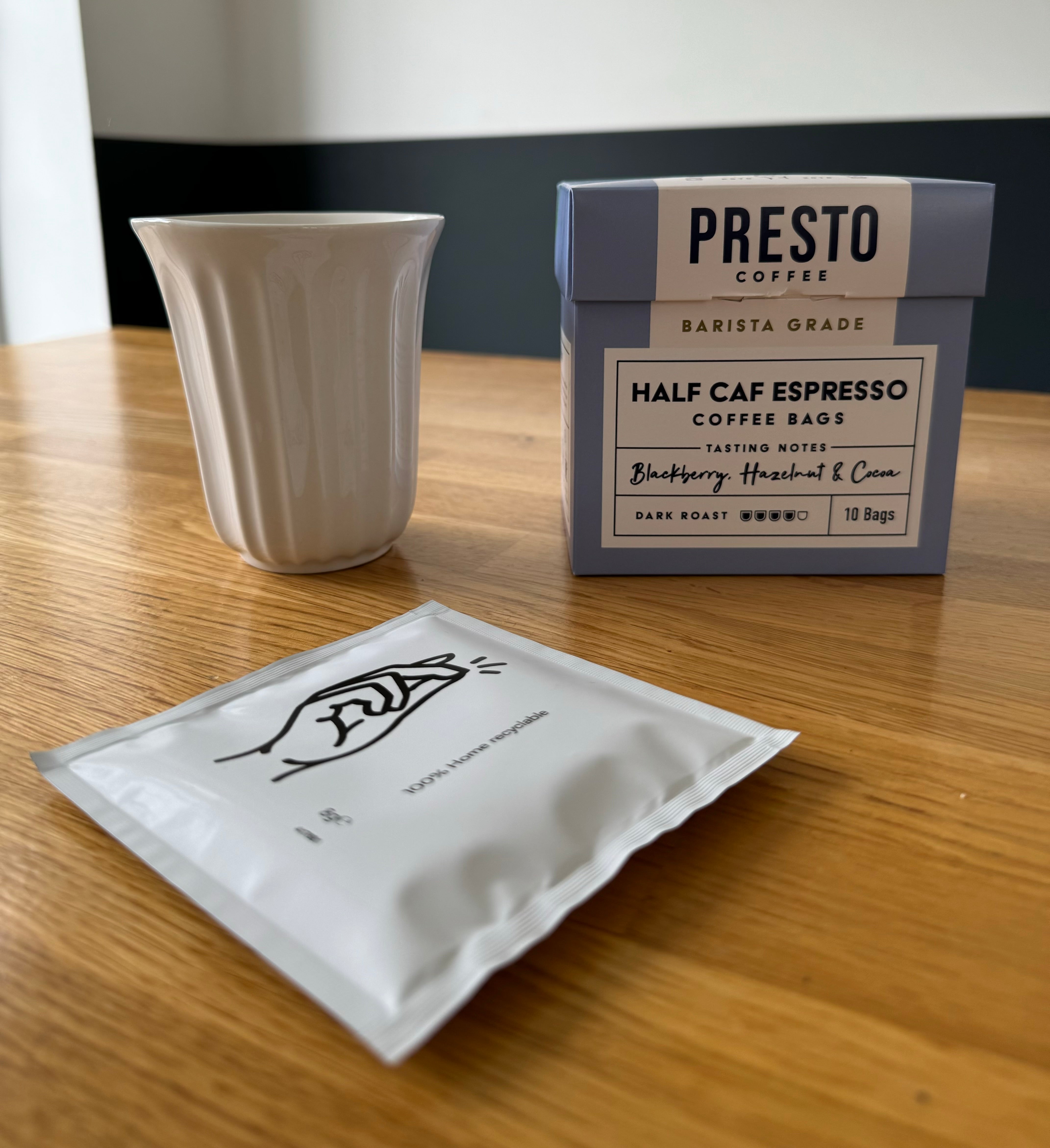 Presto Coffee Half Caf Espresso package with tasting notes of blackberry, hazelnut, and cocoa alongside a ceramic mug and a recyclable coffee bag on a wooden table.