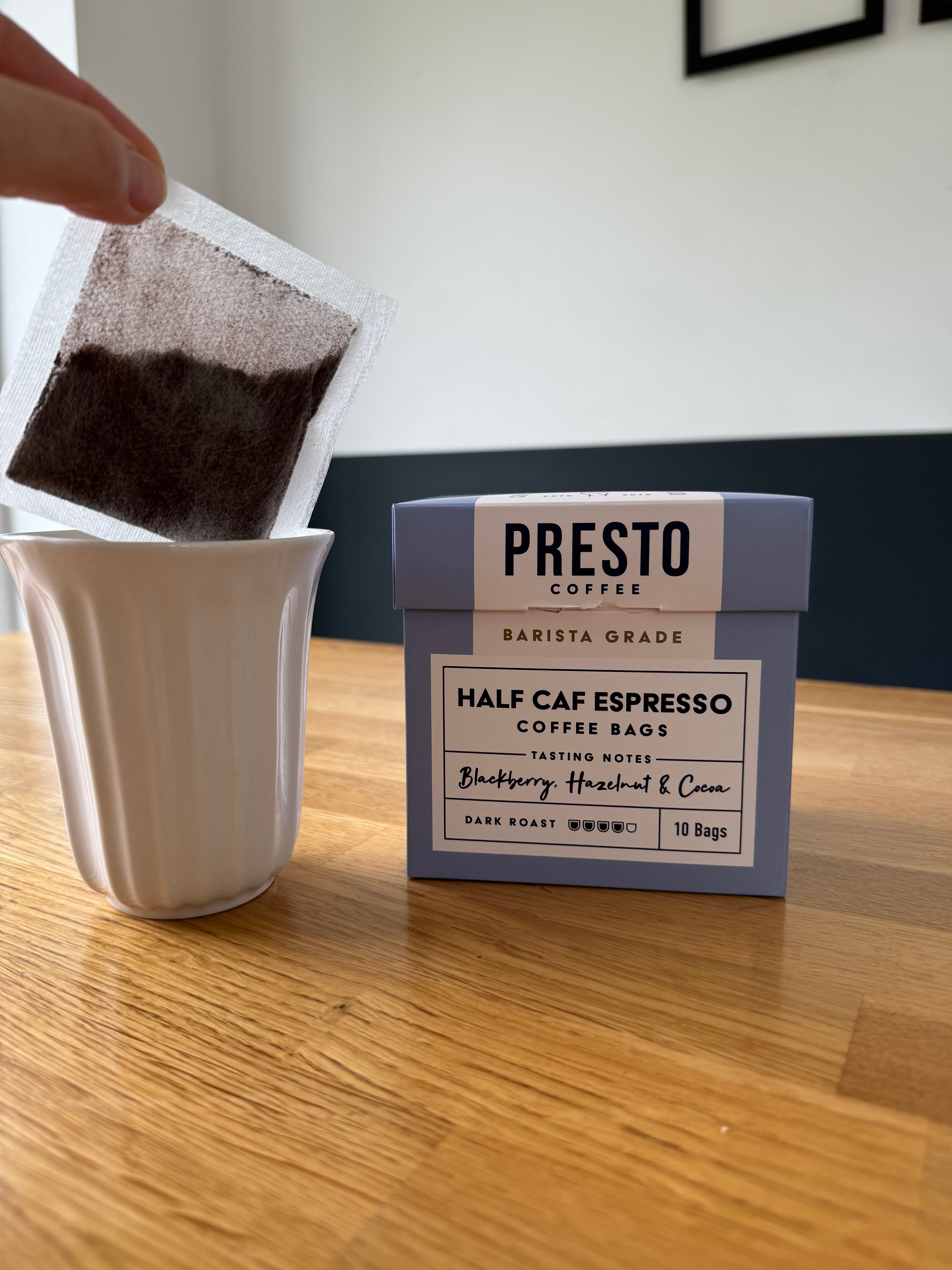 Hand holding a coffee bag over a white cup next to a box of Presto Coffee half caf espresso, dark roast, with tasting notes of blackberry, hazelnut, and cocoa on a wooden table.