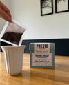 Person holding coffee bag over white cup, Presto Coffee House Decaf packaging, light roast, milk chocolate and almond notes, barista grade, on wooden table.