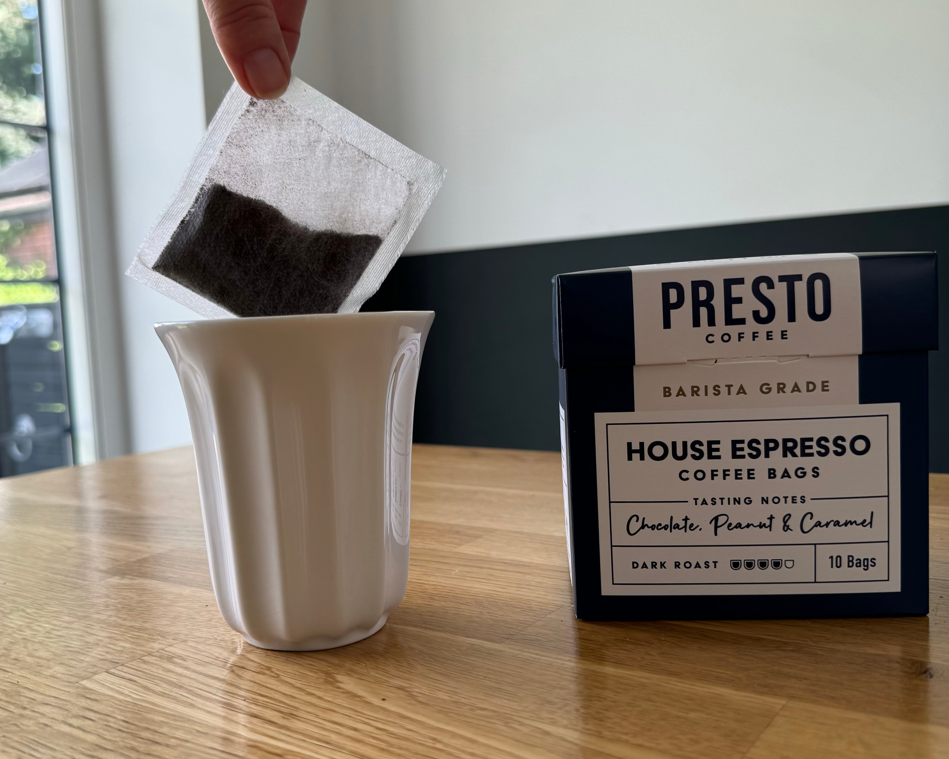 Person placing a coffee bag into a white mug next to a box of Presto Coffee House Espresso, barista grade, with tasting notes of chocolate, peanut, and caramel.