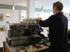 Barista preparing coffee at a professional espresso machine in a cozy cafe setting, with cups stacked on top, tools on the counter, and ambient lighting.