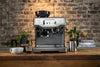 Espresso machine with various coffee drinks on a wooden table, industrial brick wall background, home coffee bar setup, modern kitchen appliance, cappuccino and latte preparation, cozy café atmosphere.